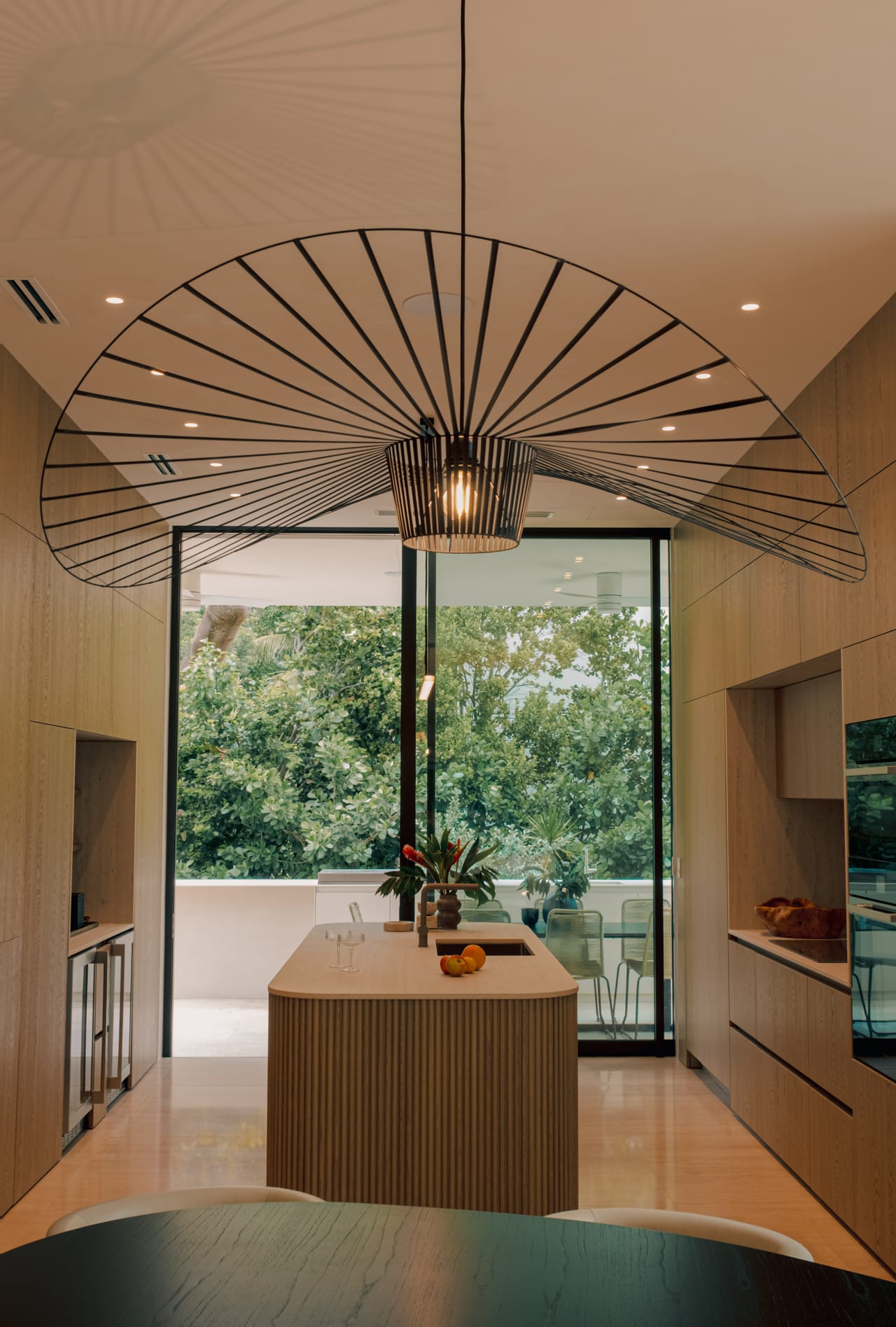 Reeded oak kitchen island beneath a sculptural pendant, open to a green terrace