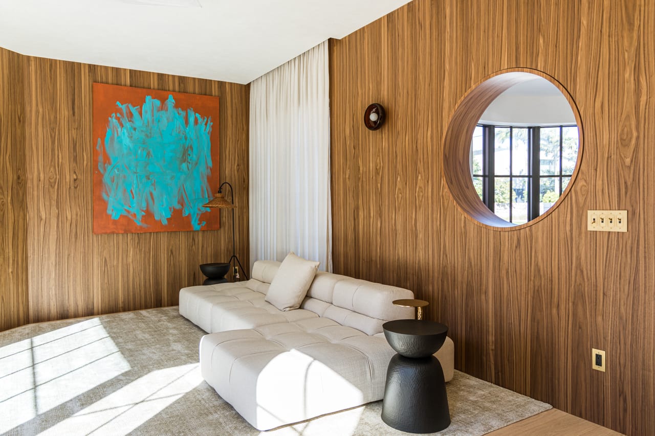 Salón revestido en nogal con ventana circular, pared curva y sofá modular color crema bajo luz rasante
